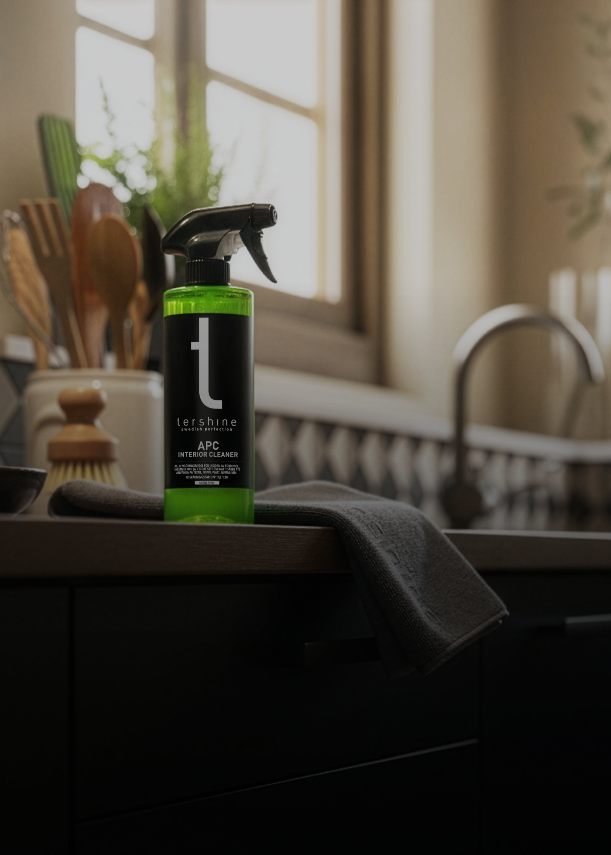 Tershine APC Interior Cleaner står på en mörk träbänk i ett svenskt kök. Varmt dagsljus från fönstret, disig bakgrund med diskho, köksredskap och kaklat stänkskydd. En grå mikrofiberduk hänger över bänkkanten. Stämningen är mörk och mysig.