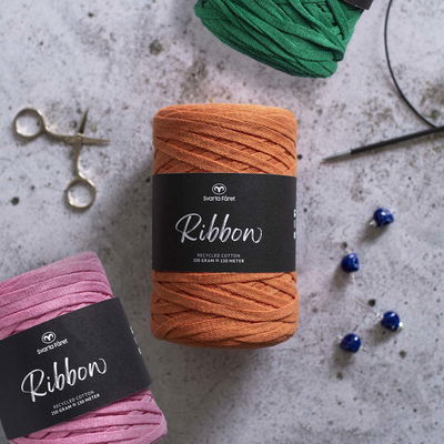 Två olika garner. orange och rosa, ur serien Ribbon från Svarta Fåret. På bilden syns även pärlor, sax och en rundsticka samt lite av ett grönt garn i ovankant