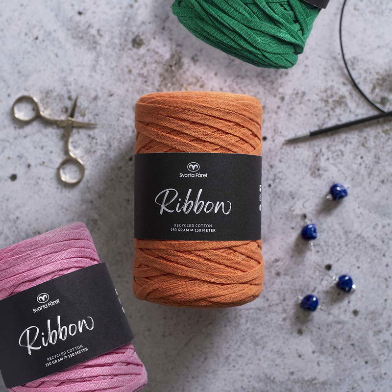 Två olika garner. orange och rosa, ur serien Ribbon från Svarta Fåret. På bilden syns även pärlor, sax och en rundsticka samt lite av ett grönt garn i ovankant