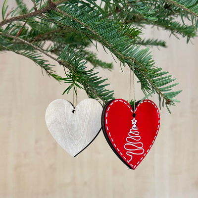Två hjärtformade julhängen i trä, ett målat rött med vit julgran och ett i naturlig plywood, hängande i grankvist