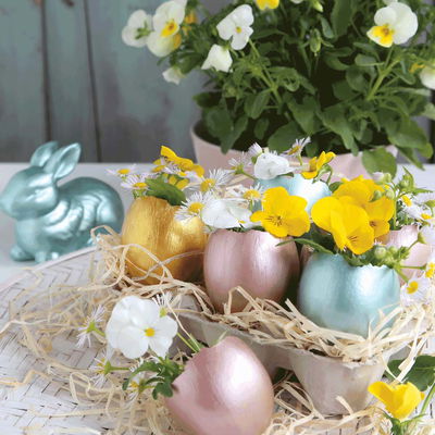 Påskägg i ett blomsterarrangemang, i bakgrunden finns även en kanin. Äggen och kaninen är målad med Marabu Colorado Gold