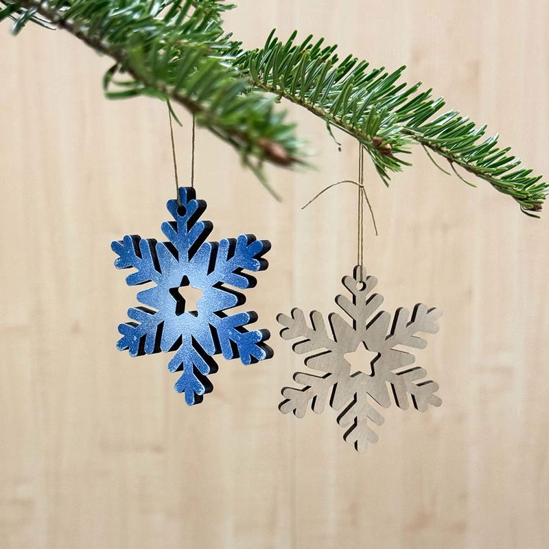 Julhänge Snöflinga, måla själv eller behåll naturligt träutseende Två trähängen i form av snöflingor hänger i en julgran, en målad i metallicblått och en i naturfärgat trä, perfekt för DIY och egen dekoration.