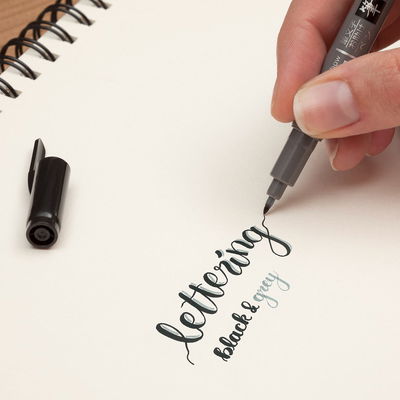 Hand som skriver “lettering black & grey” med Tombow Fudenosuke Twin, en kalligrafipenna med svart och grå penselspets