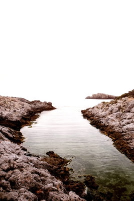 Rocky harbor vertical – motivvy. Bruna klippor ramar in ett smalt, stilla vattenparti som öppnar sig mot en ljus horisont.
