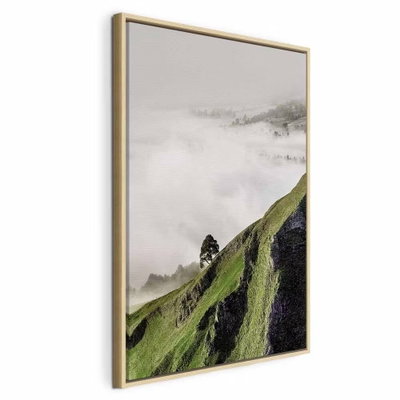 Tree above clouds vertical – naturfärgad ram. En brant grön sluttning med ett litet träd sticker upp över vita moln mot en grå horisont.