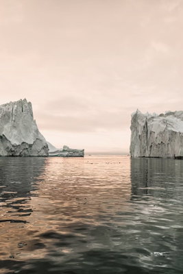 Ice statues vertical – motivvy. Två stora, ljusa isformationer speglas i stilla vatten under en blekt rosafärgad himmel.