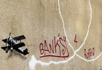 Banksy and s plane red – motivvy. Ett mörkt litet flygplan och långa vita rökspår omger röd handskriftsliknande text på en brungrå botten.