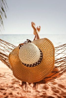 Hammock on the beach – motivvy. En stor stråhatt ligger framför uppsträckta ben i en hängmatta över en varm sandstrand.