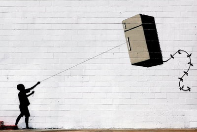 Flying fridge wide – motivvy. En svart barnsilhuett drar i ett snöre kopplat till ett svävande kylskåp framför en vit tegelvägg.
