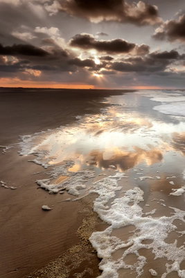 Present distraction vertical – motivvy. Vita vågor rullar in över en sandstrand under en dramatisk molnhimmel med ett smalt varmt ljus vid horisonten.
