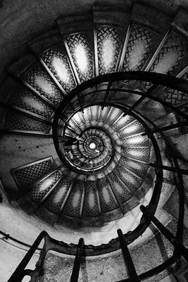 Dizziness vertical – motivvy. En mörk spiraltrappa med perforerade steg och svängda räcken bildar ett tätt, cirkelformat mönster.