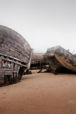 Abandoned cutters vertical – motivvy. Stora, slitna träbåtar vilar lutade på brun sand under en stor och ljusgrå himmel.
