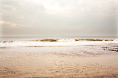 Sea after storm wide – motivvy. Små vågor möter en bred sandstrand under en stor, ljus himmel i beige och grå toner.