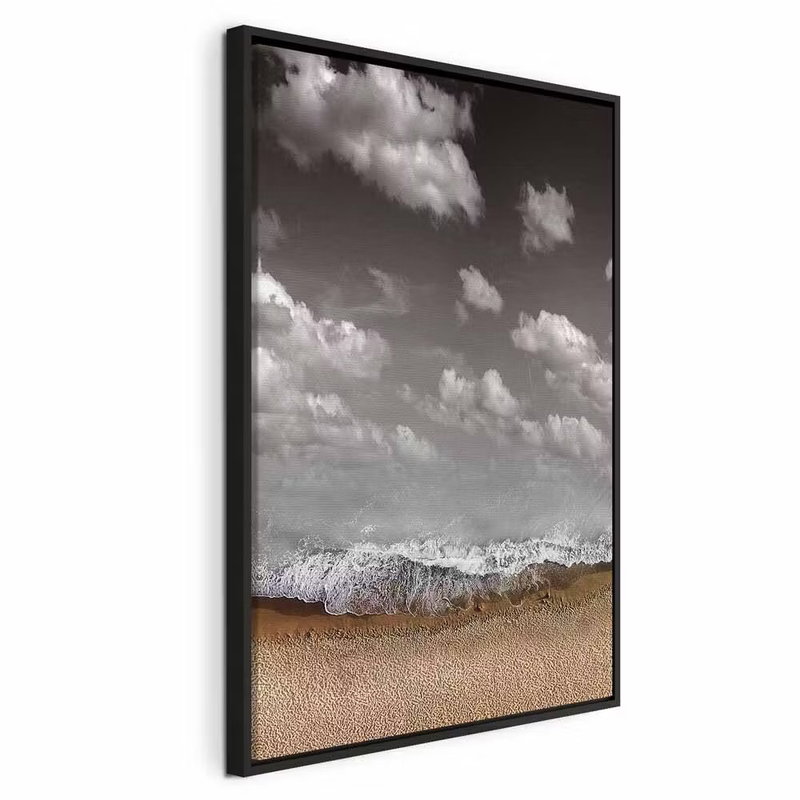 Sky wave vertical – svart ram. Ljusa moln på mörkare grå himmel möter en vit vågkant och en sandfärgad strand i en lodrät komposition.