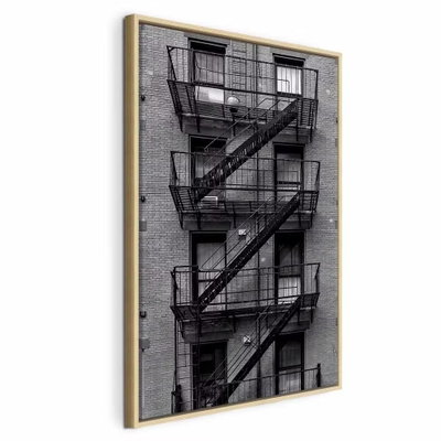 Emergency exit vertical – naturfärgad ram. Svarta räcken, brandtrappor och vilplan löper i diagonaler över en tegelklädd husfasad.