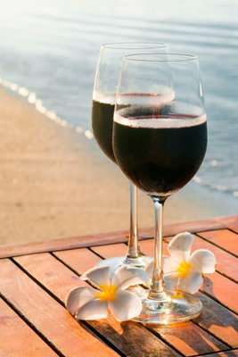 Love refreshment vertical – motivvy. Två glas med mörk dryck står på ett träbord med ljusa blommor framför en sandstrand och lugnt vatten.