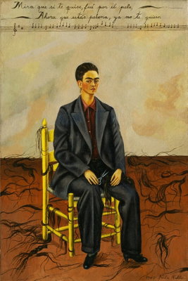 Frida kahlo self portrait – motivvy. En mörkhårig person i kostym sitter på en gul stol på sprucken mark under en ljus vägg med text.