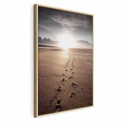 Our way vertical – naturfärgad ram. Fotspår leder genom mörk sand mot ett ljust solparti över havet under en grå himmel.