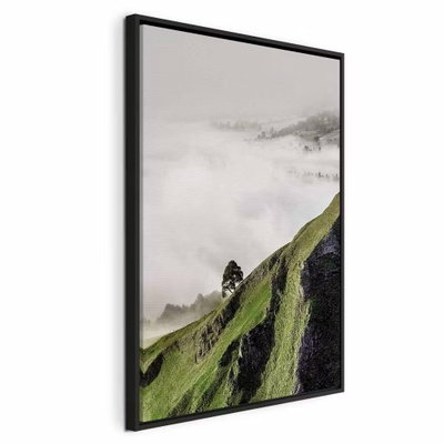 Tree above clouds vertical – svart ram. En brant grön sluttning med ett litet träd sticker upp över vita moln mot en grå horisont.
