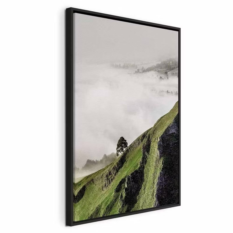 Tree above clouds vertical – svart ram. En brant grön sluttning med ett litet träd sticker upp över vita moln mot en grå horisont.