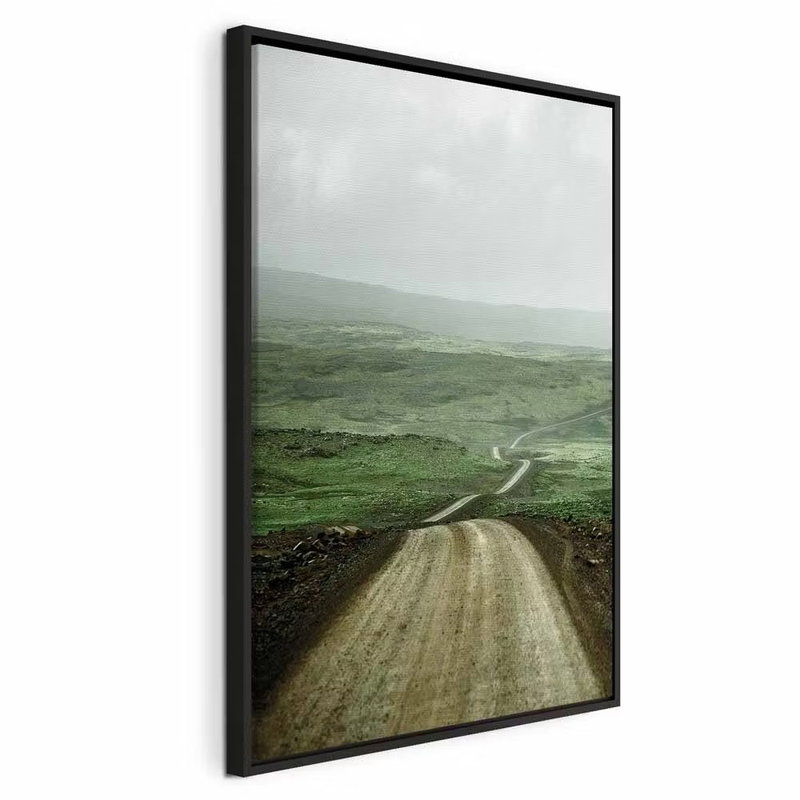 Road across the plains – svart ram. En grusväg slingrar sig genom öppna gröna slätter mot disiga kullar under en grå himmel.