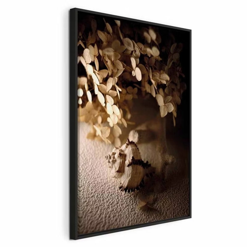 Humming seashell vertical – svart ram. En liten snäcka ligger framför ett kärl med ljusa, torra blad i varmt sidoljus.