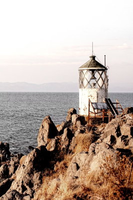 Lantern on the cliff – motivvy. En liten vit fyr står på en brant klippa vid ett lugnt hav under en ljus himmel.