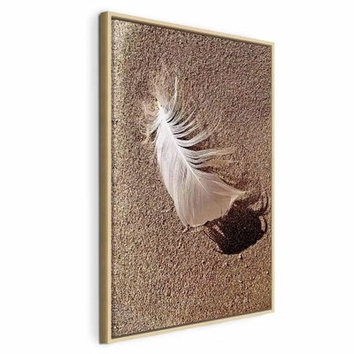 Feather on the sand vertical – naturfärgad ram. En liten vit fjäder ligger på finkornig brun sand och kastar en tydlig skugga åt sidan.
