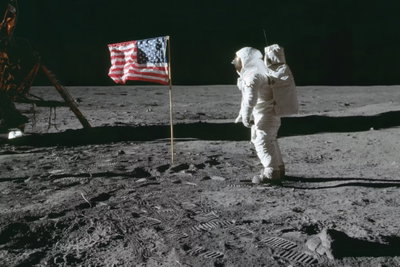 Always first wide – motivvy. En vitklädd astronaut står vid en amerikansk flagga och teknisk utrustning på en grå yta mot svart rymd.