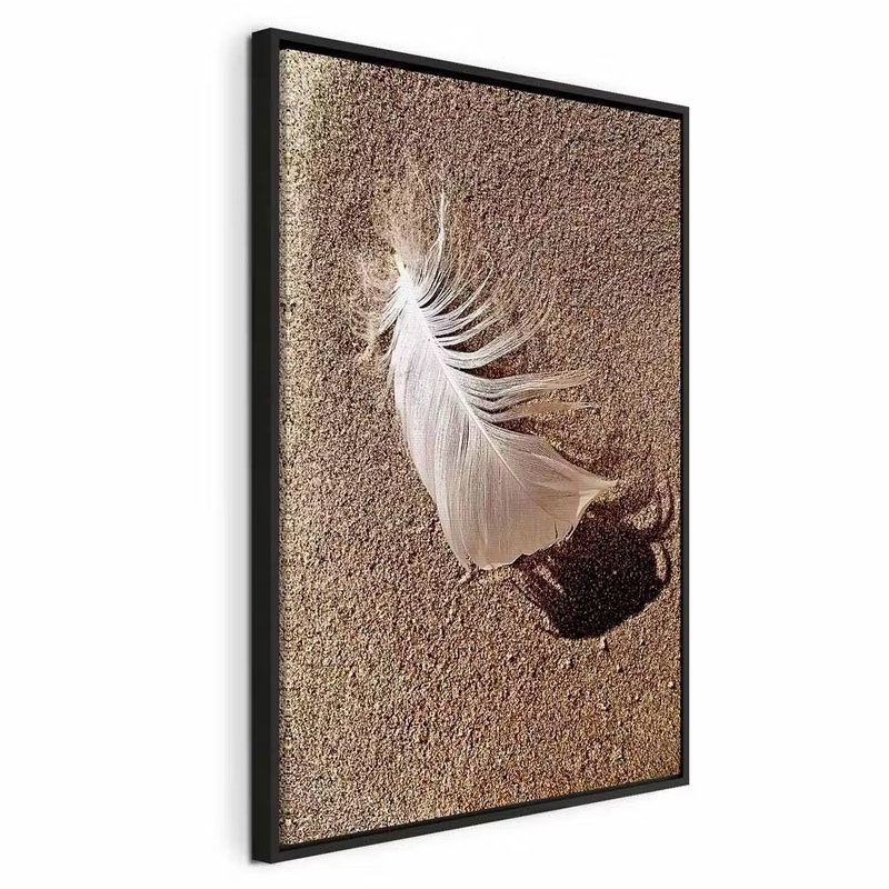 Feather on the sand vertical – svart ram. En liten vit fjäder ligger på finkornig brun sand och kastar en tydlig skugga åt sidan.