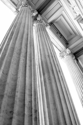 Stone columns vertical – motivvy. Höga, räfflade stenpelare ses nedifrån i svart och vitt med starka linjer mot en ljus himmel.