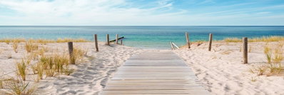 The silence of the sea – motivvy. En träspång leder genom sand och strandgräs mot ett turkost hav. Den låga horisonten sträcker ut landskapet.