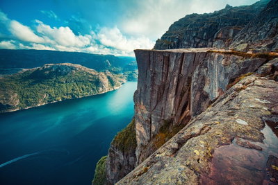 Rock city vertical – motivvy. En hög, rak klippkant reser sig över djupt blått vatten i ett bergigt landskap under molnig himmel.