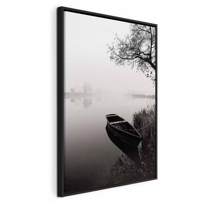 Haven under tree vertical – svart ram. En mörk träbåt ligger vid en strand under ett utsträckt träd i ett dimmigt svartvitt landskap.