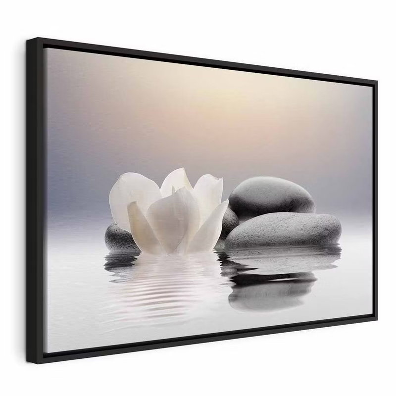 Garden of calm – svart ram. En vit blomma ligger intill staplade, rundade grå stenar som speglas i en stilla vattenyta.