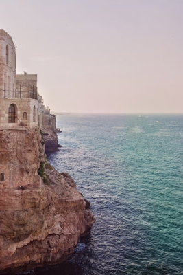 Turquoise coast vertical – motivvy. Stenbyggnader ligger på en klippkant ovanför turkost vatten, under en ljus himmel.
