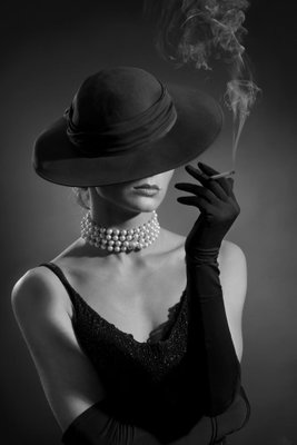 Black elegance vertical – motivvy. En kvinna i mörk hatt, handskar och pärlhalsband håller en cigarett med en liten rökstrimma mot svart.