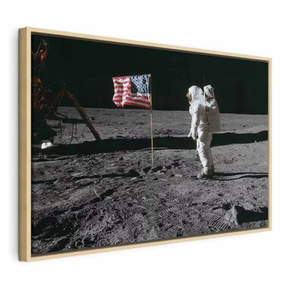 Always first wide – naturfärgad ram. En vitklädd astronaut står vid en amerikansk flagga och teknisk utrustning på en grå yta mot svart rymd.