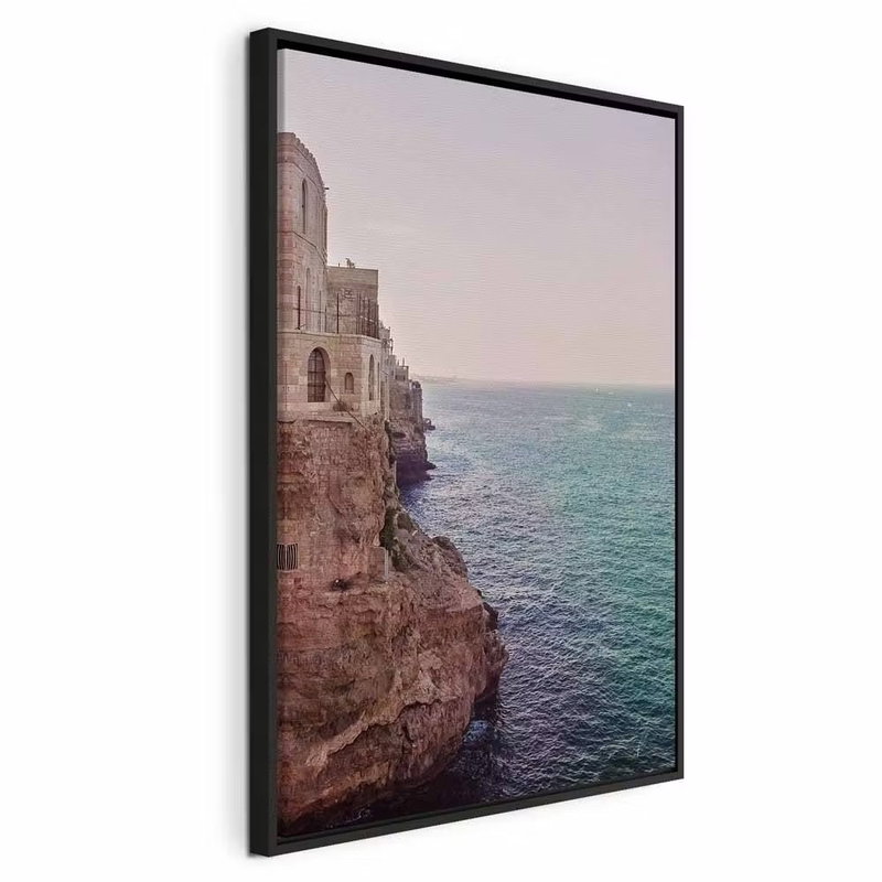 Turquoise coast vertical – svart ram. Stenbyggnader ligger på en klippkant ovanför turkost vatten, under en ljus himmel.
