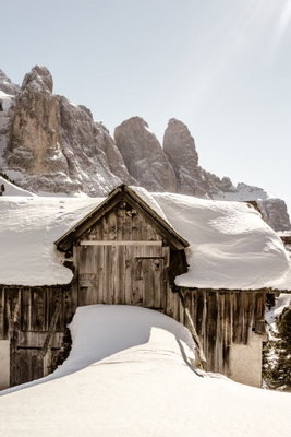 Winter in the dolomites – motivvy. En mörk trästuga ligger inbäddad i snö nedanför höga, klippiga bergstoppar.