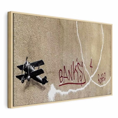 Banksy and s plane red – naturfärgad ram. Ett mörkt litet flygplan och långa vita rökspår omger röd handskriftsliknande text på en brungrå botten.